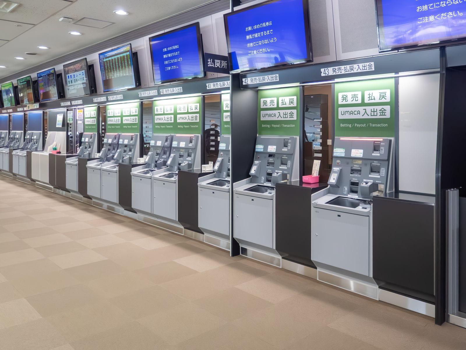 A long row of combined betting and payout machines with green signs inside a Tokyo Racecourse stand.