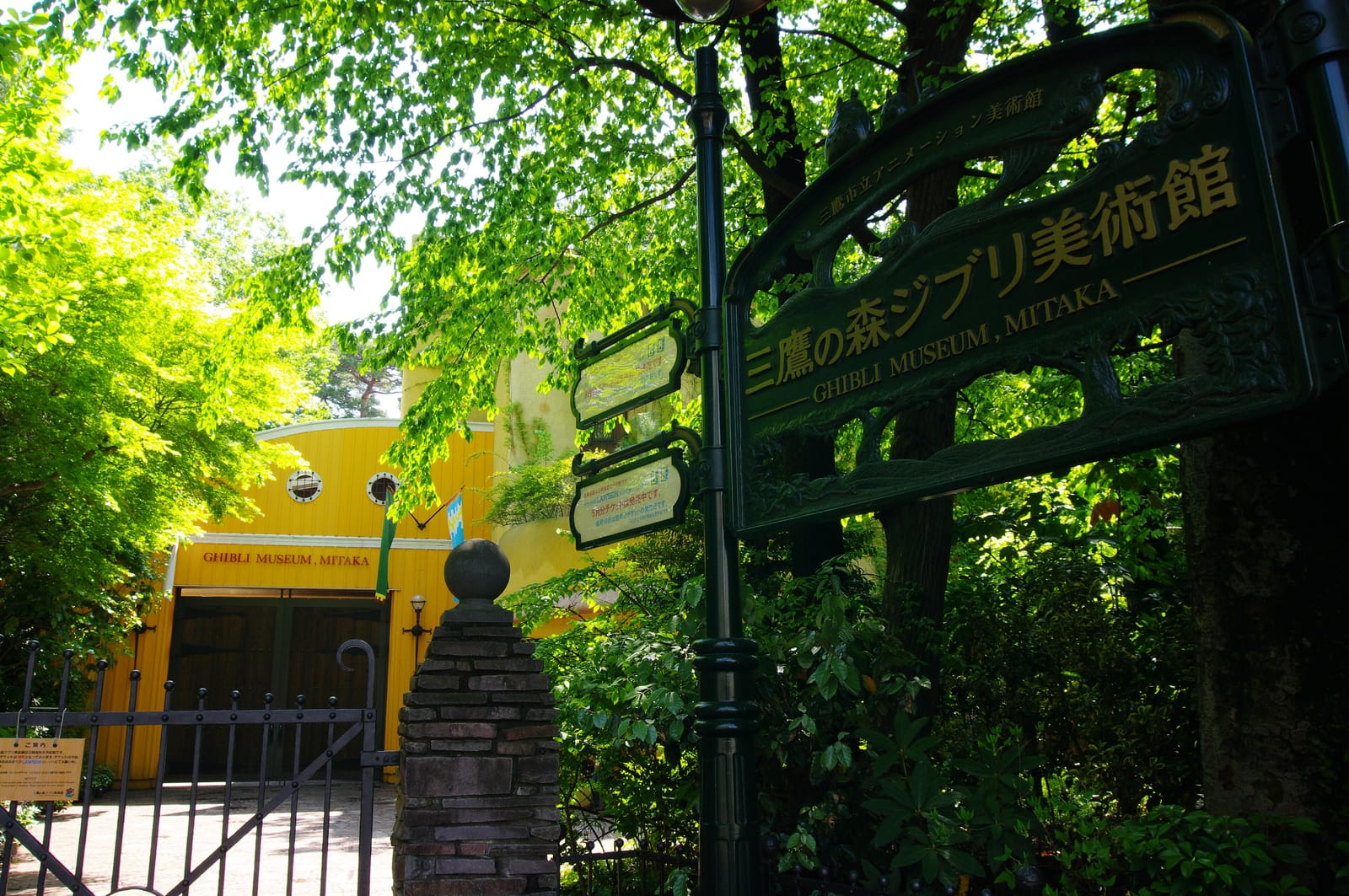 The Ghibli Museum, Mitaka sign and gate, with the yellow entrance building behind fresh green trees